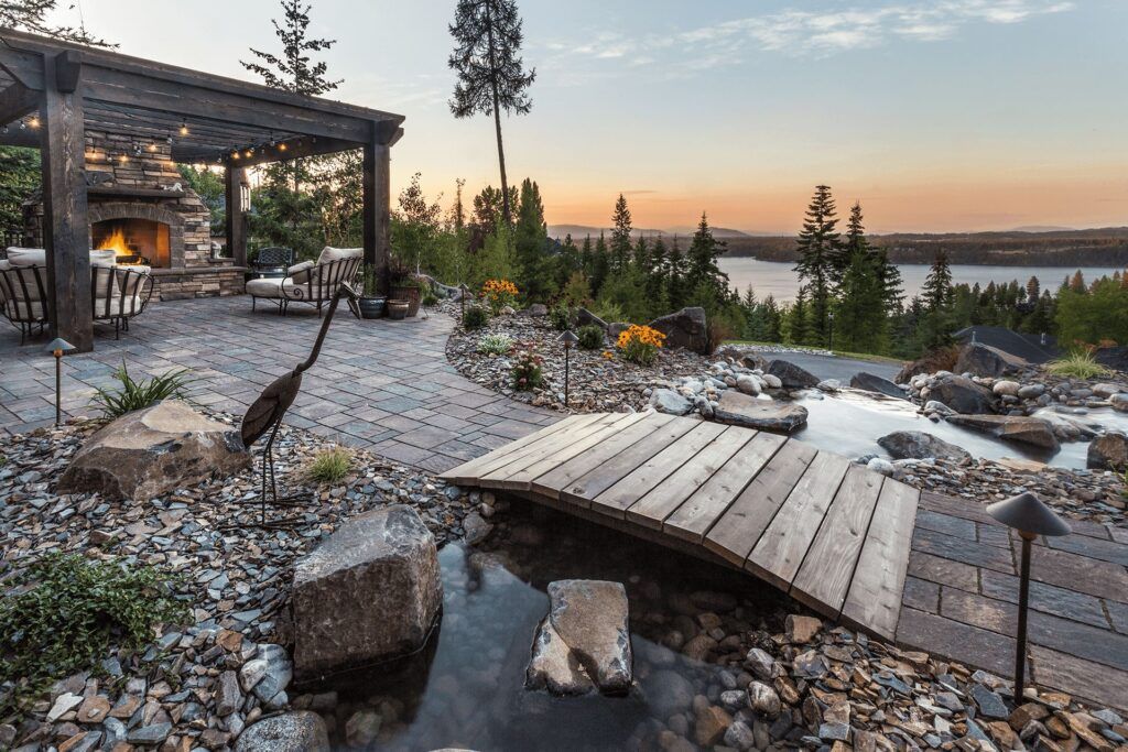 A beautifully landscaped backyard by Copper Creek Landscaping features a stone patio with a fireplace under a pergola, a wooden footbridge over a stream, vibrant flower gardens, and scenic lake views framed by trees at sunset.