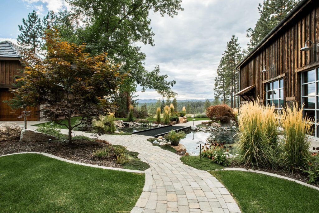 A curved stone pathway by Copper Creek Landscaping leads through a landscaped garden with a pond, tall grasses, shrubs, rocks, and trees beside rustic wooden buildings under a partly cloudy sky.