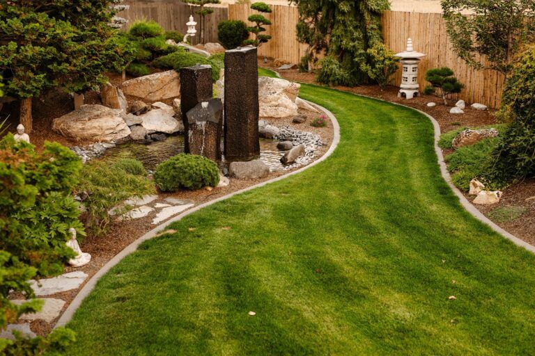 A beautifully landscaped garden by Copper Creek Landscaping features a curved grass pathway, stone border, water feature with upright stones, rocks, shrubs, and a white ornamental lantern near a wooden fence.